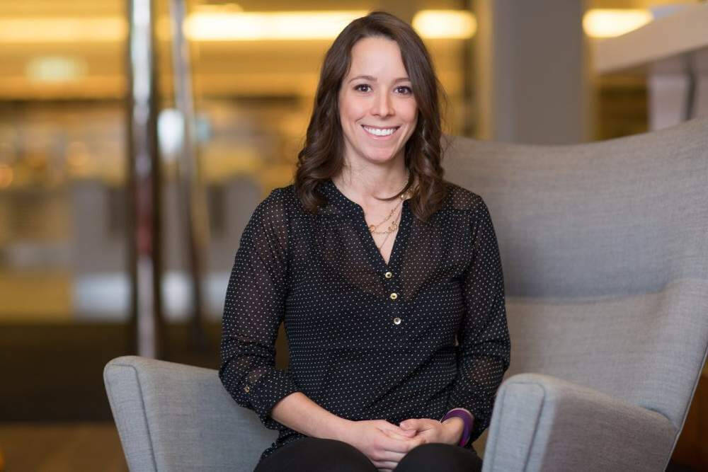 A woman sitting on a gray chair, smiling, wearing a black blouse with small white dots, in a warmly lit indoor setting.